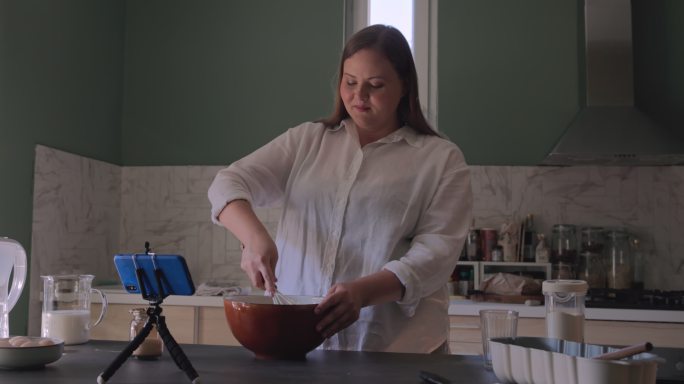 快乐超重的女面包师在厨房里做面团，在三脚架上用手机按照食谱做