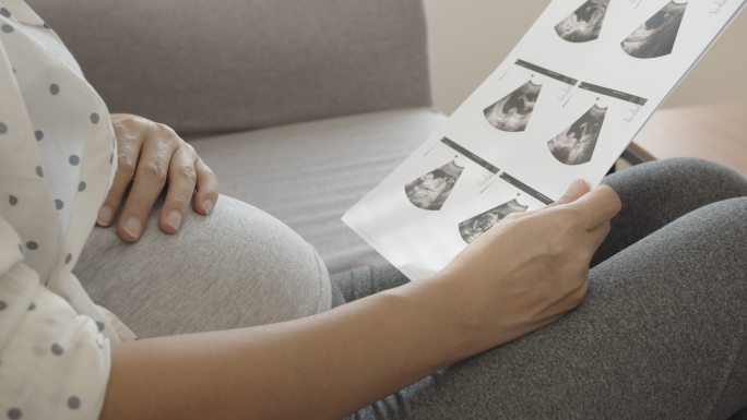 一名亚洲孕妇坐在沙发上看超声波图像并触摸腹部。