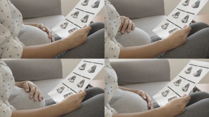 一名亚洲孕妇坐在沙发上看超声波图像并触摸腹部。