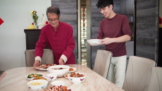 亚裔中国老人带着儿子从厨房里拿食物，在中国新年期间摆在餐桌上