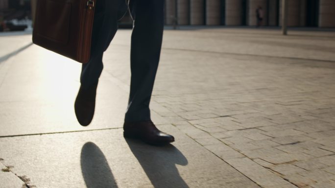 一个穿着棕色皮鞋的商人的脚，在一个美丽的夜晚穿过一条铺好的街道，我们可以看到他的腿和棕色笔记本电脑包