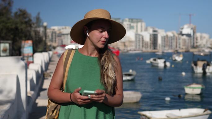 一个戴着漂亮帽子的游客走在码头上。她欣赏港口和船只的景色