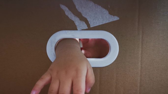 亚裔中国男婴从纸箱上的椭圆洞里偷看隐藏在纸箱里，用窗框阴影向外看