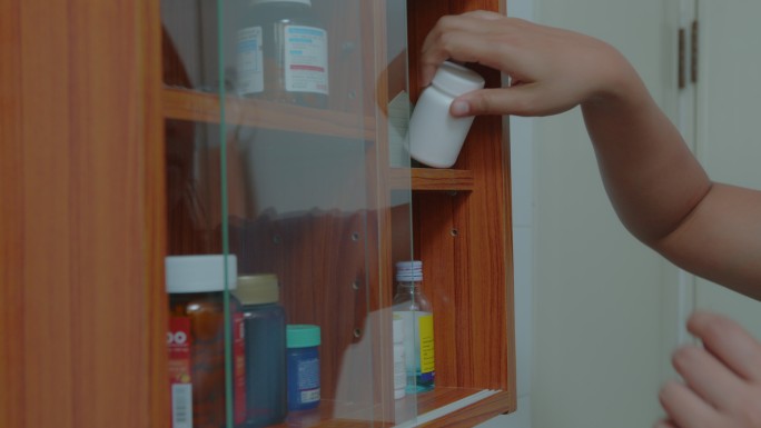 一个亚洲女人打开浴室药柜的门，拿出一瓶药。健康和医学概念。