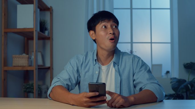 一名年轻的亚洲男子晚上坐在家里客厅的办公桌上，用手机在线购物。
