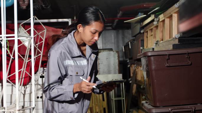 一名20多岁的亚裔妇女穿着灰色制服，在仓库储藏室清点物品。维修服务