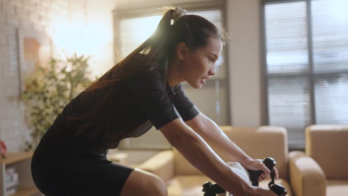 一个骑自行车的亚裔女子。她正在家里锻炼。通过在教练机上骑自行车和玩在线自行车游戏