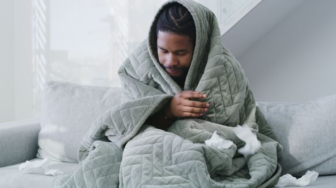 一个年轻人在家里得了流感，躺在床上。感觉不舒服的人，用毯子盖住，用纸巾擤鼻涕