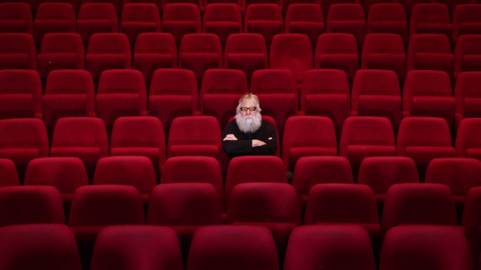 一个留着白胡子的男人坐在空荡荡的电影院或剧院里
