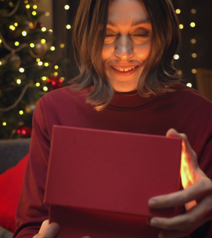 一个棕色头发的年轻漂亮女人打开一个礼物盒的特写镜头，背景是圣诞灯