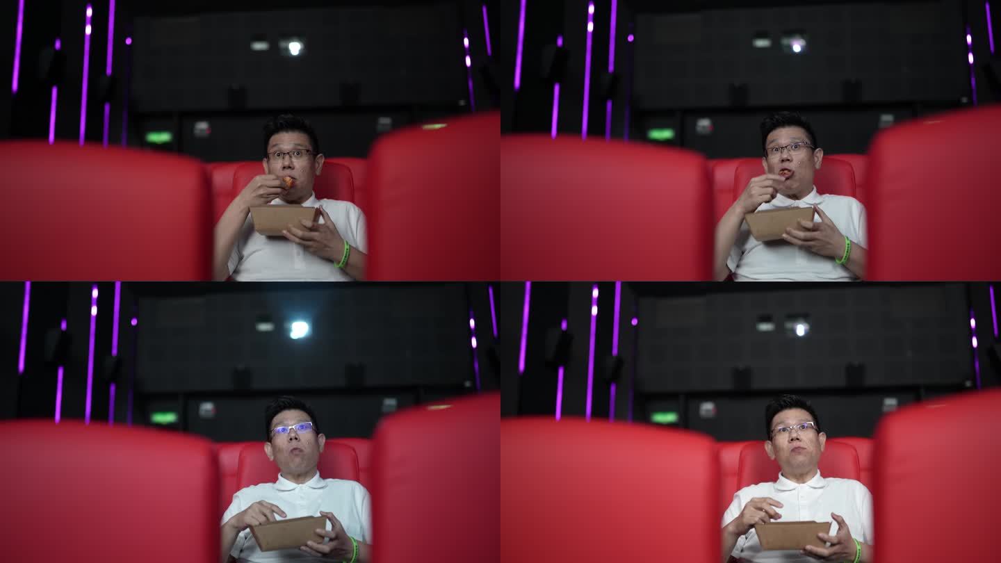 一名亚裔中国男子坐在空荡荡的电影院红色座位上吃东西，欣赏电影。