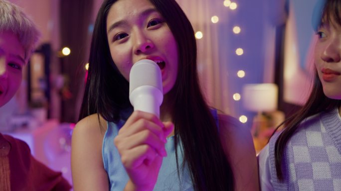 年轻的亚洲女性朋友唱卡拉OK的特写镜头，晚上在多彩的家庭聚会上玩得很开心。