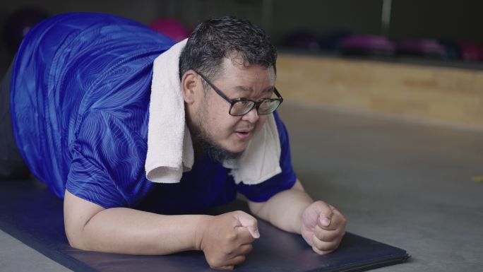 在室内健身中心，一个精疲力竭的超重年轻人正努力在垫子上做平板运动，气喘吁吁。