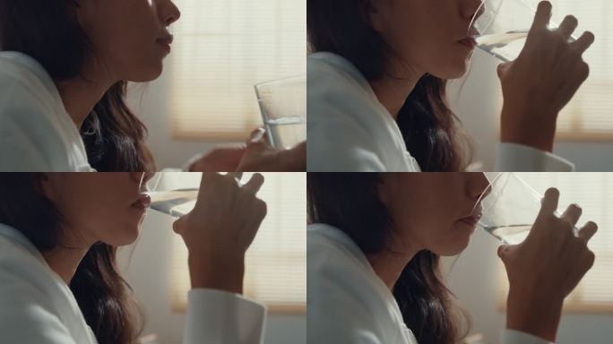 特写镜头：一位生病的年轻亚洲妇女坐在客厅沙发上的毯子下，喝着处方药和水，头痛或抑郁。