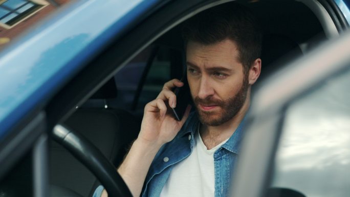 一位留着胡子的欧洲男子坐在驾驶座上，开着车门，在电话里讲话