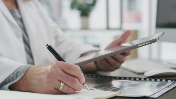 一名无法辨认的医生在医院办公室使用数字平板电脑时在笔记本上写字