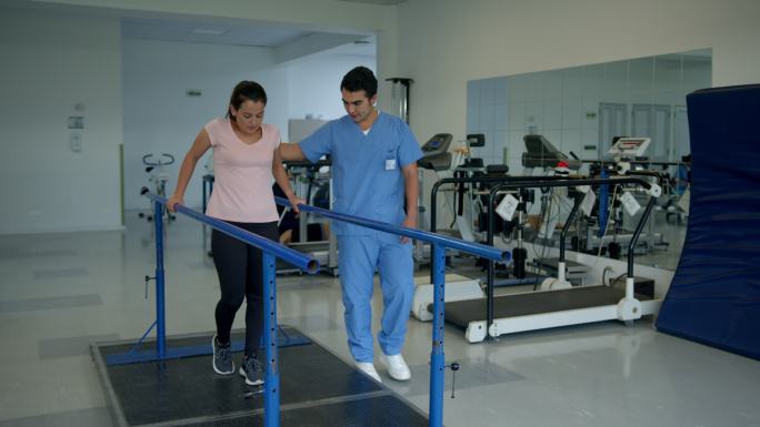 在物理治疗期间，年轻女性患者靠着双杠用力行走，而男性治疗师在她旁边行走