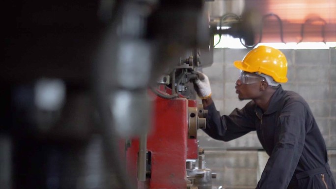 在工厂特写镜头中，一名在控制面板上工作的非洲裔美国人与一台工业机器一起工作。在工业机械中，男性工程师