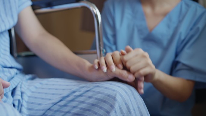 亚洲护士的特写手握住坐在轮椅上的男性患者的手，在辅助生活和癌症斗争中给予支持和同情。
