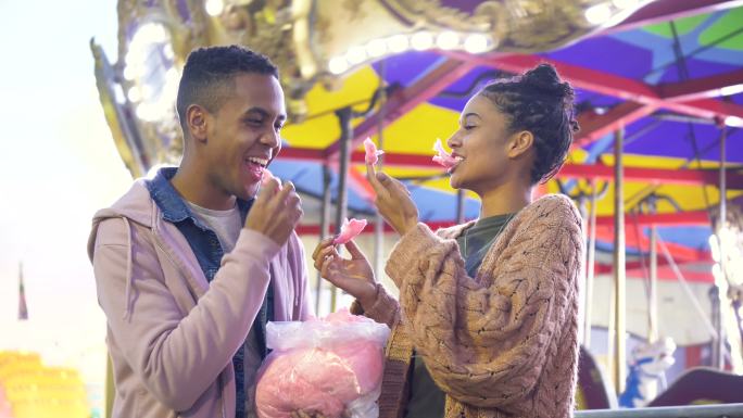 一对年轻的黑人夫妇在狂欢节上吃着棉花糖，笑着