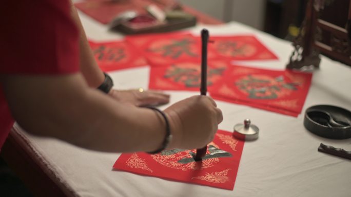 亚洲华人男性为即将到来的中国新年庆祝活动练习中国书法的手，在红纸上书写，以繁荣和良好的措辞装饰家居