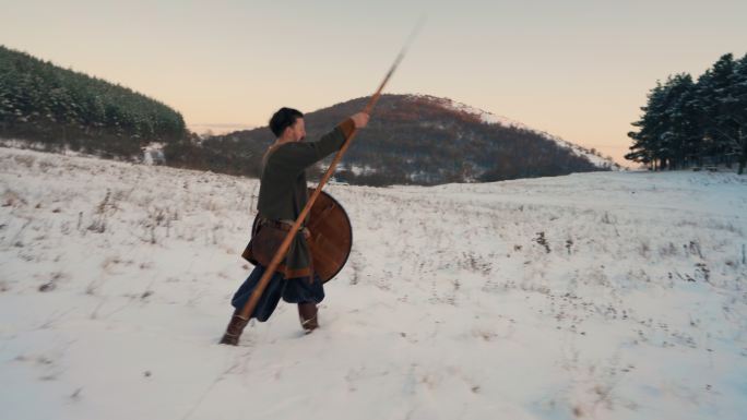 一位中世纪战士在日落时手持盾牌和长矛行走在白雪覆盖的草地上