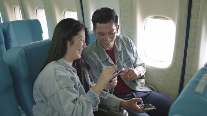一对穿着牛仔衬衫的亚洲旅行者夫妇坐在飞机的窗户旁，在假期旅行中使用智能手机