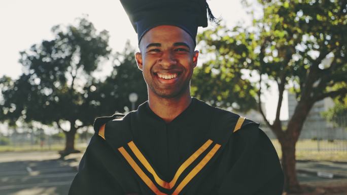 一位年轻的男学生的肖像，他交叉着双臂站在大学外面，看起来很开心。骄傲、微笑的男人准备毕业并庆祝获得学