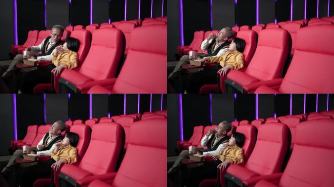 亚裔中国爷爷和孙女坐在空荡荡的电影院里的红色座位上，一边吃一边看电影