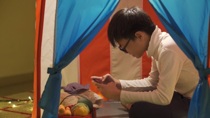 一个亚裔中国少年在家里一个舒适的照明帐篷里使用手机