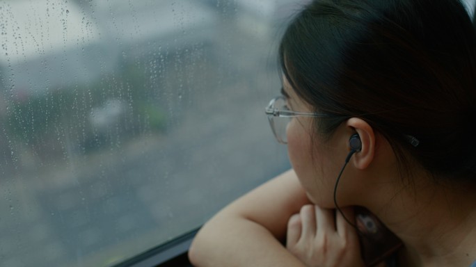 悲伤的女人坐在窗前，看着雨，听着音乐