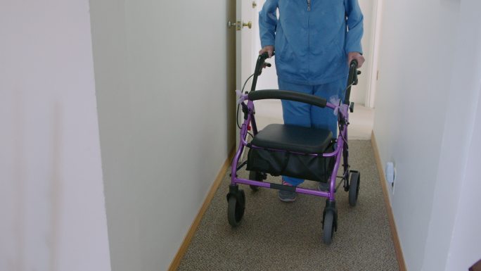 手持式拍摄的一名独立的老年妇女带着移动助行器穿过家中的走廊