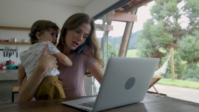 美丽的女人在家里工作，带着蹒跚学步的孩子，用笔记本电脑打电话