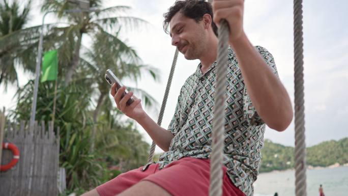 在海滩上，年轻人在荡秋千绳上荡秋千，同时通过手机进行视频通话