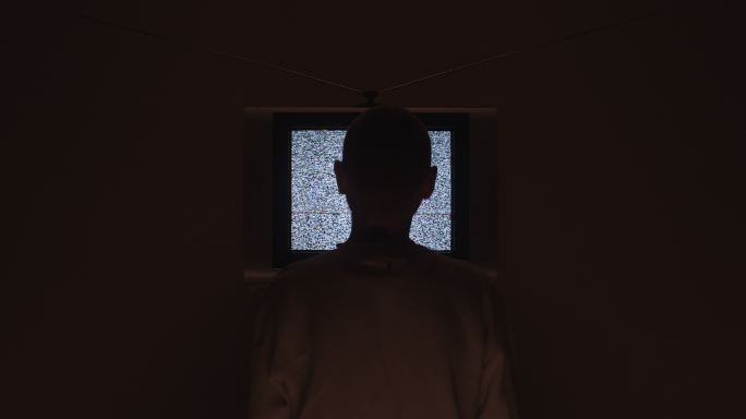 一名男子进入画面，坐在电视机前，在一间黑暗的房间里，画面中央有电视静电噪音