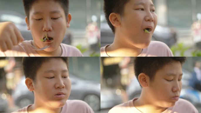 泰国华欣，一名亚洲男孩在露天街头食品餐厅与家人一起吃早餐时，正在吃蔬菜叶子。