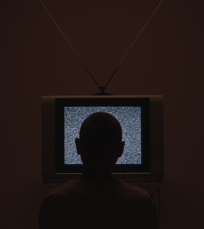 一个男子进入房间坐在电视机前的垂直视频，在黑暗的光线下，画面中间有电视静态噪音
