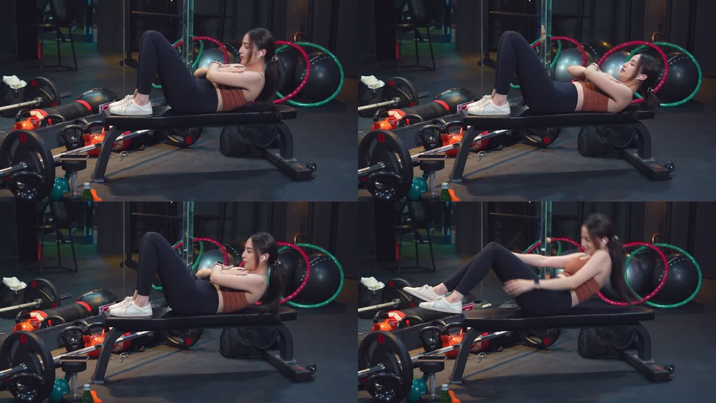 一名亚洲年轻女运动员坐在运动椅上做仰卧起坐，在健身房腹痛。一个健身女性正在做腹肌仰卧起坐。健身房的腹