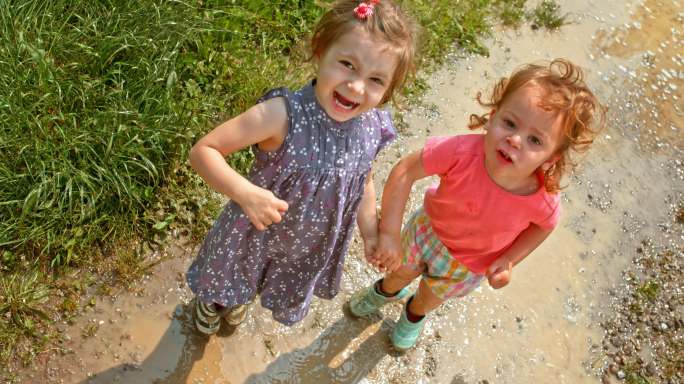 斯洛·莫：两个小女孩手牵着手在水坑里跳跃，对着镜头微笑