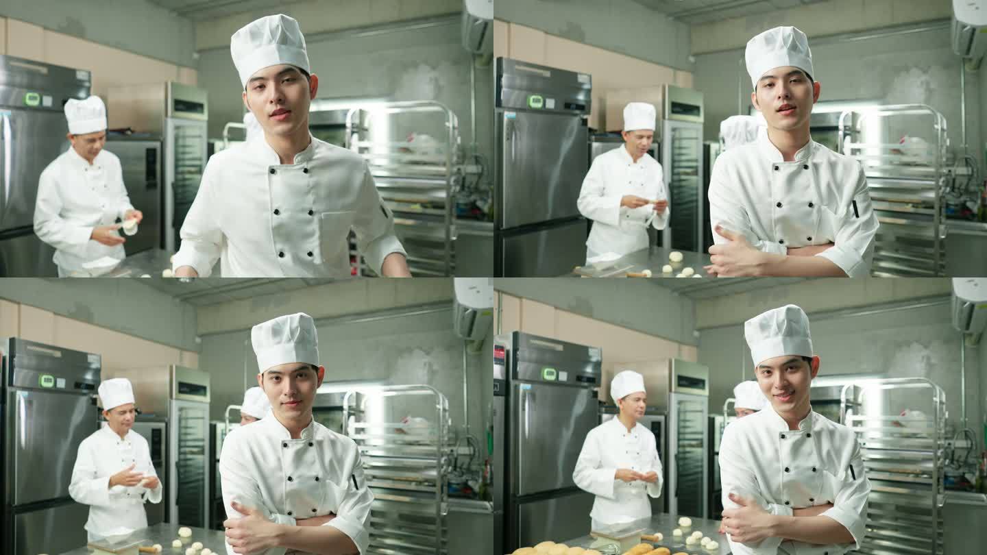 肖像：快乐的Asain英俊的年轻男子厨师站着，微笑着交叉双臂，靠在厨房柜台上，以其他厨师为背景，在不