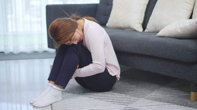 一名亚洲妇女在家中躺着或坐在沙发上胃痛，期间感到疼痛
