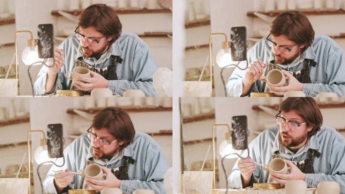 一名男性陶工在陶艺工作室用雕刻工具雕刻粘土时，正在手机上录制视频。内容创作者和技艺精湛的工匠在他的艺