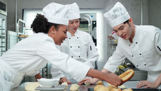 穿着白色厨师制服的亚洲和非洲年轻厨师一起工作，制作面包和面包，在烤箱中进行打样和烘焙之前进行揉捏和成