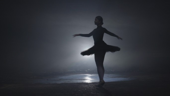 身着黑色芭蕾舞短裙的芭蕾舞演员在舞台上舞动着神奇的蓝光和烟雾。穿着芭蕾舞鞋的年轻迷人舞者的剪影在黑暗