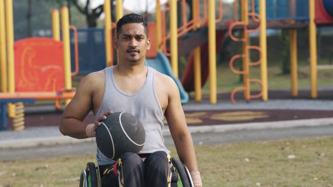 坐在轮椅上拿着健身球的亚洲印度残疾人男子在公园晨练中自信地以冷静的态度看着摄像机