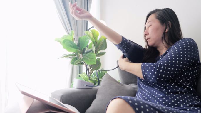 亚洲女性进行血压测试，坐在家里靠窗的沙发上，在网站上查看数字表寻求建议，健康的生活方式理念。