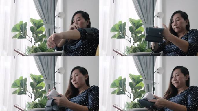 亚洲女性进行血压测试，坐在家里靠窗的沙发上，在网站上查看数字表寻求建议，健康的生活方式理念。