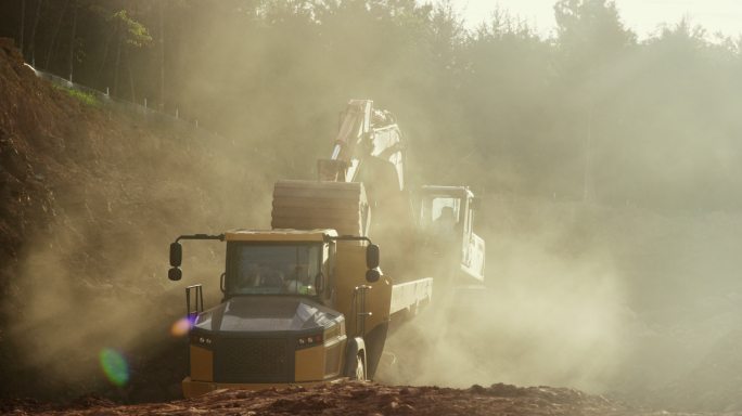 挖掘机在多尘施工现场向自卸卡车后部倾倒泥土的慢动作广角镜头（铰接式自卸卡车）