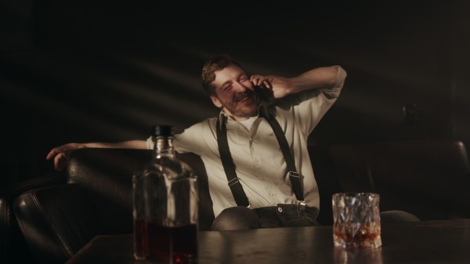 一个穿着衬衫和背带裤的醉汉坐在沙发上，喝着威士忌，他接了电话，先高兴地说了起来，然后开始讲述他的悲伤