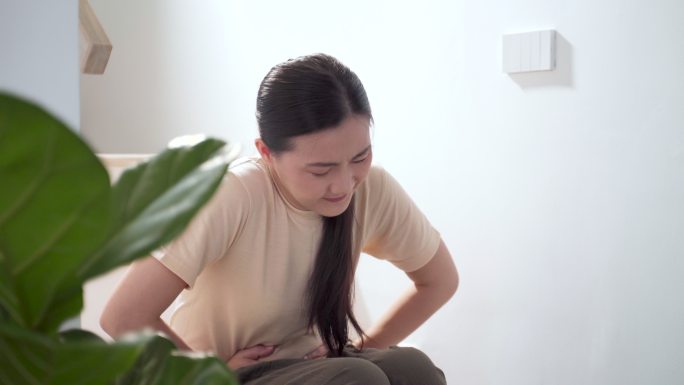 一位亚洲妇女坐在家里的楼梯上，胃痛得很厉害。